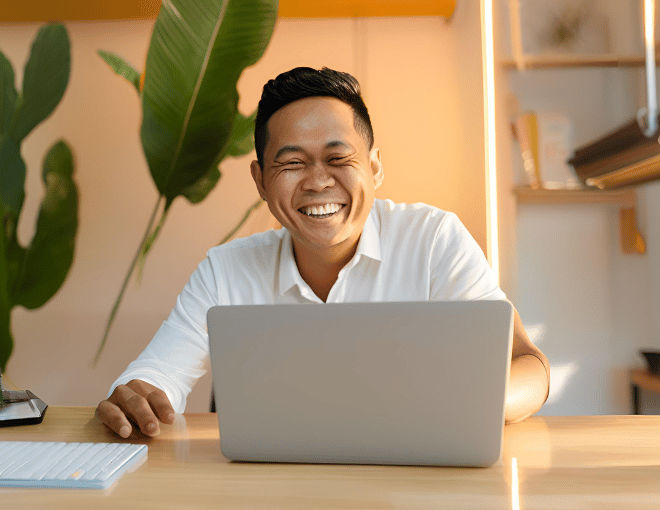 An Indonesian man sits at his desk, fingers poised over the laptop keyboard, light reflecting off the glossy surface, his eyes filled with joy.